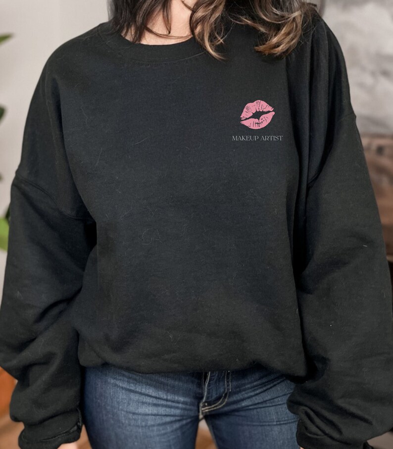 Peut inclure: Sweat-shirt noir avec un imprim&eacute; de rouge &agrave; l&egrave;vres rose et le texte "MAKEUP ARTIST" sur le devant.