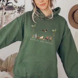 K&ouml;nnte beinhalten: Ein gr&uuml;ner Kapuzenpullover mit einer Tasche und einem grafischen Aufdruck von Tieren und B&auml;umen. Der Text "Respect the Locals" ist unter dem Aufdruck gedruckt.