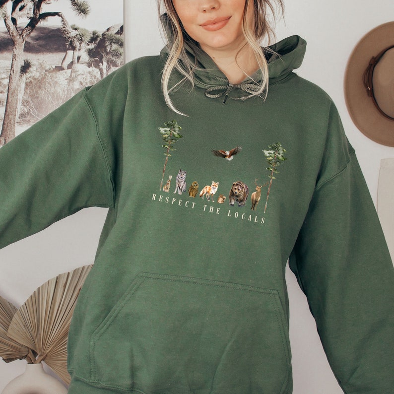 K&ouml;nnte beinhalten: Ein gr&uuml;ner Kapuzenpullover mit einer Tasche und einem grafischen Aufdruck von Tieren und B&auml;umen. Der Text "Respect the Locals" ist unter dem Aufdruck gedruckt.