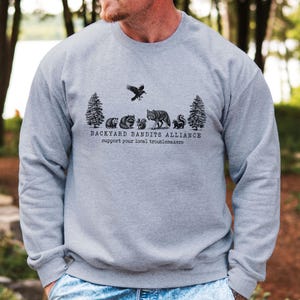 May include: A gray sweatshirt with a black graphic print of a forest scene with animals and the text "Backyard Bandits Alliance support your local troublemakers".
