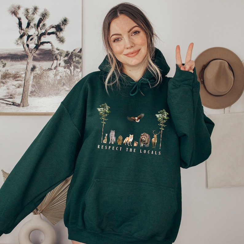 K&ouml;nnte beinhalten: Ein dunkelgr&uuml;ner Kapuzenpullover mit einem wei&szlig;en Grafikdruck einer Reihe von Tieren in einer Waldlandschaft. Der Text "Respect the Locals" ist unter den Tieren gedruckt.