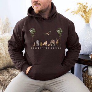 K&ouml;nnte beinhalten: Ein braunes Hoodie mit einem Aufdruck von Tieren und B&auml;umen. Der Text "Respect the Locals" ist unter dem Aufdruck gedruckt.