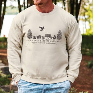 May include: A beige sweatshirt with a graphic of a forest scene with animals and the text "Backyard Bandits Alliance support your local troublemakers".