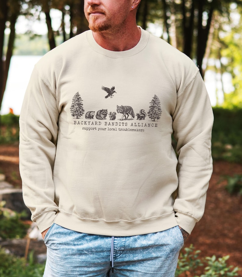 May include: A beige sweatshirt with a graphic of a forest scene with animals and the text "Backyard Bandits Alliance support your local troublemakers".