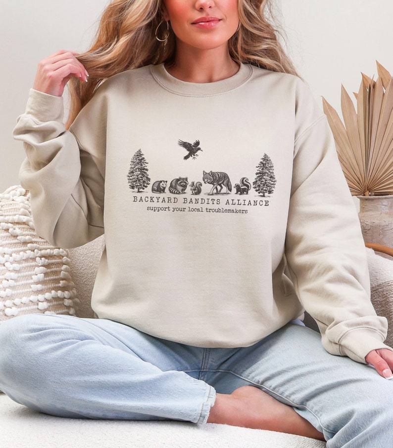 May include: A light beige sweatshirt with a graphic print of a forest scene with animals and the text "BACKYARD BANDITS ALLIANCE support your local troublemakers".
