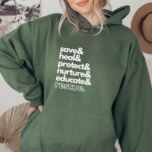 Peut inclure: Un sweat &agrave; capuche vert fonc&eacute; avec une poche avant et une capuche &agrave; cordon. Le sweat &agrave; capuche pr&eacute;sente un texte blanc qui se lit comme suit : "save & heal & protect & nurture & educate & rescue." Un chapeau beige est accroch&eacute; au mur en arri&egrave;re-plan.
