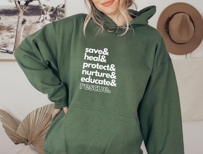 Peut inclure: Un sweat &agrave; capuche vert fonc&eacute; avec une poche avant et une capuche &agrave; cordon. Le sweat &agrave; capuche pr&eacute;sente un texte blanc qui se lit comme suit : "save & heal & protect & nurture & educate & rescue." Un chapeau beige est accroch&eacute; au mur en arri&egrave;re-plan.