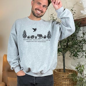 May include: A gray sweatshirt with a black graphic print of a crow, trees, and animals. The text "BACKYARD BANDITS ALLIANCE" and "support your local troublemakers" is printed below the graphic.