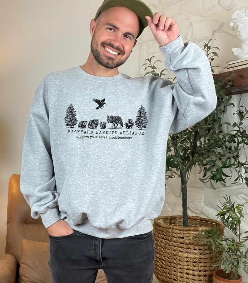 May include: A gray sweatshirt with a black graphic print of a crow, trees, and animals. The text "BACKYARD BANDITS ALLIANCE" and "support your local troublemakers" is printed below the graphic.