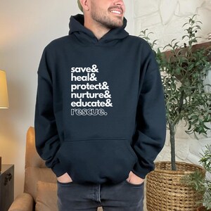 Peut inclure: Sweat-shirt &agrave; capuche noir avec texte blanc : "save & heal & protect & nurture & educate & rescue." Le sweat-shirt a une poche avant et une capuche &agrave; cordon. La personne porte un jean fonc&eacute;.