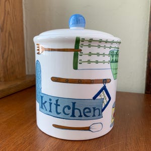 May include: White ceramic kitchen canister with a lid and a blue knob. The canister is decorated with kitchen utensils and the word "kitchen" in blue. The design includes a blue oven mitt, a green pot holder, and a wooden spoon.