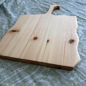 May include: A light-coloured wooden serving board with a natural grain pattern and visible knots. The board features a rectangular body with wavy, organic edges and a single handle extending from the top centre, crafted from smooth, unfinished wood.