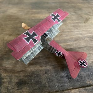 May include: A miniature biplane model in burgundy with silver wings and black crosses. The toy airplane has a propeller and a tail fin. The model is set against a wooden background.