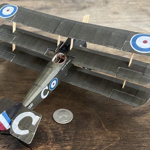 May include: A detailed model of a triplane aircraft, featuring three wings and a dark green color scheme. The model includes roundels with blue and red accents, and the text "Black Maria" is visible. A quarter coin is next to the model.