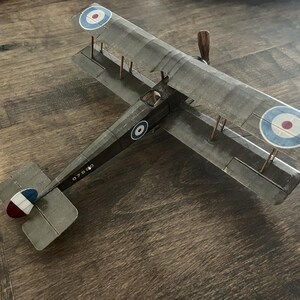 May include: A gray biplane model with roundels on the wings and fuselage. The model has a wooden propeller and a tail fin with red, white, and blue accents. The wings are connected by struts.