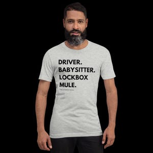 May include: A light gray t-shirt with the words "DRIVER. BABYSITTER. LOCKBOX MULE." printed in black. The text is stacked vertically. Below the text is the phrase "THE LOCKBOX MULE." in a smaller font.
