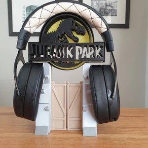 May include: A 3D printed headset stand featuring the Jurassic Park logo. The stand is white and gray with a black headset resting on it.