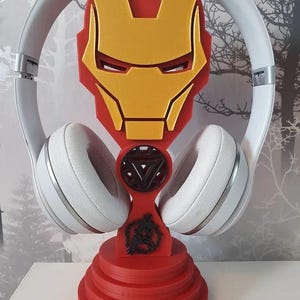 May include: A red and yellow Iron Man-themed headphone stand. The stand features the Iron Man mask design and a circular base. White headphones are placed on the stand. The stand is made of plastic.