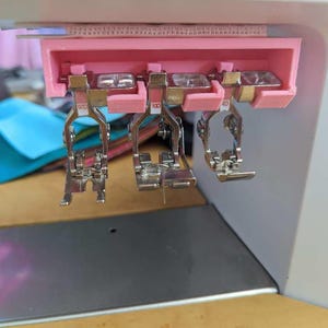 Puede incluir: Tres pies de máquina de coser están unidos a un soporte rosa. Los pies están etiquetados con "90", "100" y "40".