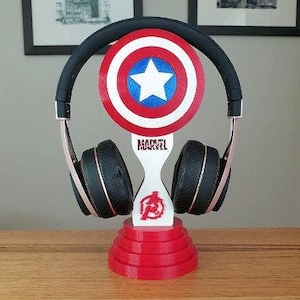 May include: A red, white, and blue Captain America shield-shaped headphone stand with the word "MARVEL" printed on the front. The stand is holding a pair of black headphones.