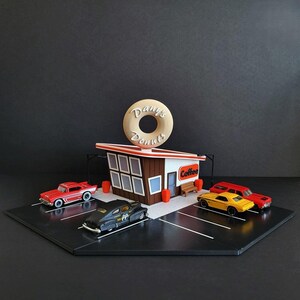 May include: A miniature model of a donut shop with a red and white building, a brown roof, and a large donut sign that says "Danny's Donuts". There are four miniature cars parked in front of the shop, including a red car, a black car, and two yellow cars. The shop has a sign that says "Coffee".