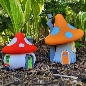 Könnte beinhalten: Zwei Miniatur-Pilzhäuser, eines rot mit weißen Punkten und eines orange mit blauen Punkten, werden auf einem Bett aus braunem Mulch präsentiert. Die Häuser sind aus Keramik und haben eine märchenhafte, fantasievolle Ästhetik.