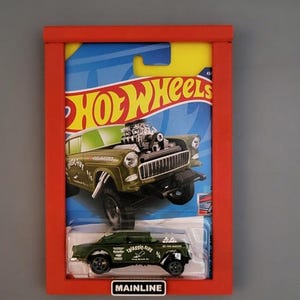 May include: A framed Hot Wheels die-cast car in a red frame. The car is olive green with black wheels and the text "Triassic-Five". The packaging features a larger image of the same car. The word "Mainline" is visible.