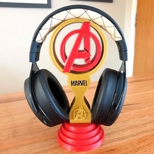 May include: A 3D printed headphone stand featuring the Avengers logo in red and gold with the word "MARVEL" printed below. The stand is made of a red and gold plastic and has a black headphone resting on it.
