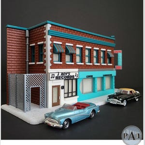 May include: A miniature model of a brick building with a turquoise awning and a sign that reads "Roy's Records". There are two vintage cars parked in front of the building, one blue and one black.