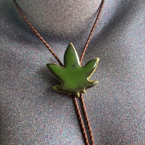 May include: A bolo tie featuring a glossy, olive-green ceramic pendant shaped like a five-pointed leaf. The pendant is centered on a braided brown leather cord that loops around the neck, providing a secure and adjustable fit for the wearer.