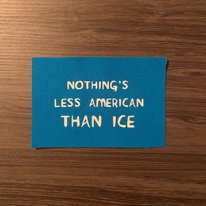 May include: A rectangular, turquoise fabric patch with the phrase "NOTHING'S LESS AMERICAN THAN ICE" printed in white, centered on the patch. The patch is set against a brown wooden surface.
