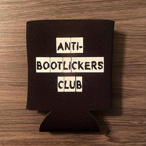 May include: Black beverage insulator with the text "ANTI-BOOTLICKERS CLUB" in white block letters. The insulator is made of a thick, textured material and has a cutout at the bottom. The background is a wooden surface.