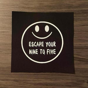 May include: A black square patch featuring a white smiley face and the text "ESCAPE YOUR NINE TO FIVE". The design is simple and bold, perfect for adding a touch of personality to clothing or accessories.