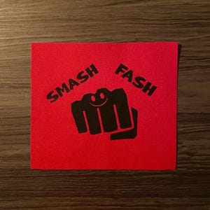 May include: A square, red patch with the words "SMASH" and "FASH" printed in black. A black fist graphic with a smiling face is centered between the words. The patch is likely for clothing or accessories.