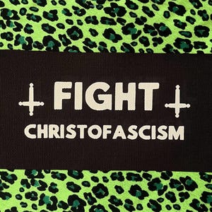 May include: A rectangular black fabric patch featuring white embroidered text that reads FIGHT CHRISTOFASCISM. The design includes a white cross symbol on the left and an inverted cross symbol on the right, centered on a bright neon green leopard print.