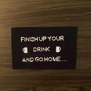 May include: Black rectangular patch with white text that reads "FINISH UP YOUR DRINK AND GO HOME...". The text is centered and includes two small mug icons. The patch is on a wooden surface.