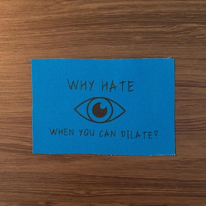 May include: A blue rectangular patch with the text "WHY HATE" above a brown eye graphic, and "WHEN YOU CAN DILATE?" below. The patch is on a wooden surface.