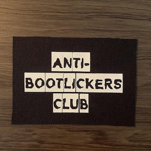 May include: Black rectangular patch with the words "ANTI-BOOTLICKERS CLUB" in white block letters. The patch is made of a textured fabric and is likely designed to be sewn or ironed onto clothing or accessories.