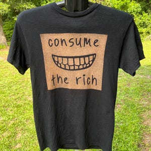 May include: A black short-sleeved t-shirt featuring a large, textured, tan-colored square graphic on the chest. The graphic displays a black line drawing of a wide, toothy grin centered between the black text that reads consume the rich.