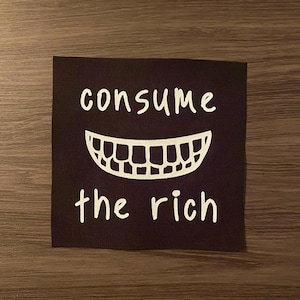 May include: Black square patch with the words "consume the rich" in white, above a white smiling mouth graphic. The patch is on a wooden surface.