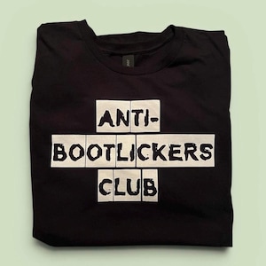 May include: Black t-shirt with the words "ANTI-BOOTLICKERS CLUB" in white block letters. The text is arranged in a cross shape. The shirt is folded neatly.