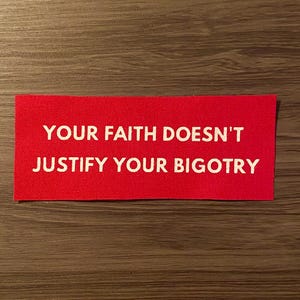 May include: A rectangular red patch with the white text: "YOUR FAITH DOESN'T JUSTIFY YOUR BIGOTRY." The patch is set against a wooden background. The text is centered and in a sans-serif font.