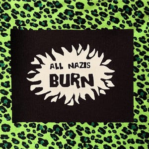 May include: A rectangular black fabric patch featuring a white, jagged, sunburst-shaped graphic in the center. The text "ALL NAZIS" is printed in black above the word "BURN," which is also printed in black, centered within the white graphic shape.