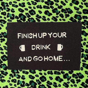 May include: A rectangular black fabric patch featuring white embroidered text that reads FINISH UP YOUR DRINK AND GO HOME with two small white mug icons flanking the word DRINK. The patch has a visible woven texture throughout the surface.
