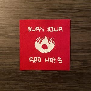 May include: A square, red fabric patch with white text and a graphic. The text reads "BURN YOUR RED HATS". The graphic is a stylized flame with a hat shape in the center. The patch is on a dark brown wooden surface.