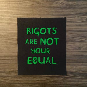 May include: Black rectangular patch with the text "BIGOTS ARE NOT YOUR EQUAL" in bright green, set against a dark wood grain background. The patch is made of a textured fabric.