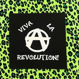 May include: A black square patch with the words "VIVA LA REVOLUTION!" in white, along with a white anarchy symbol. The background is a neon green leopard print.