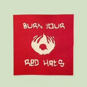 May include: A square, red fabric patch with white text and a graphic. The text reads "BURN YOUR RED HATS" with a flame-like graphic in the center. The patch has a textured appearance.