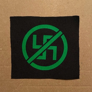 May include: A black square patch with a green symbol of a swastika inside a circle with a diagonal line through it. The patch is made of a woven fabric.