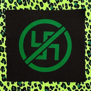 May include: A square black fabric patch featuring a green circular emblem with a swastika symbol crossed out by a diagonal line. The patch is centered against a bright neon green and black leopard print background with a textured fabric weave.